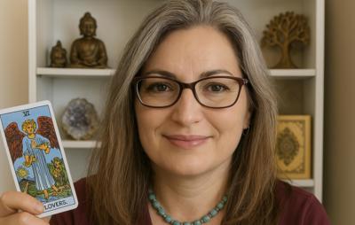 Een vrouw met halflang, grijzend haar en een warme glimlach houdt een tarotkaart omhoog. Ze draagt een bril en een turquoise kralenketting. Op de acht